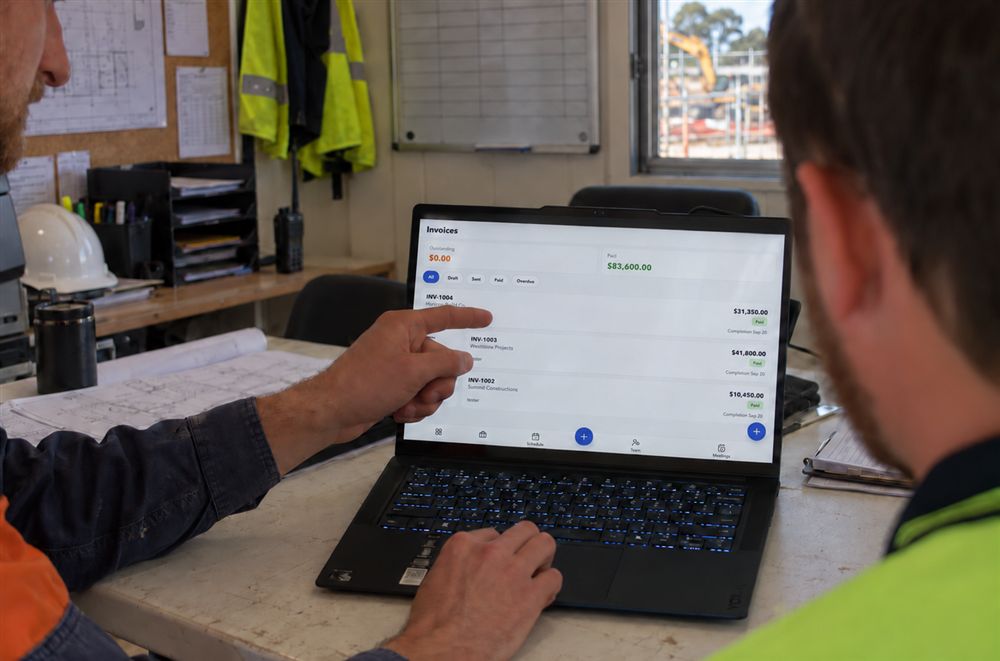 Two tradies reviewing paid invoices on a laptop in the site office, using the Voloflo invoicing screen
